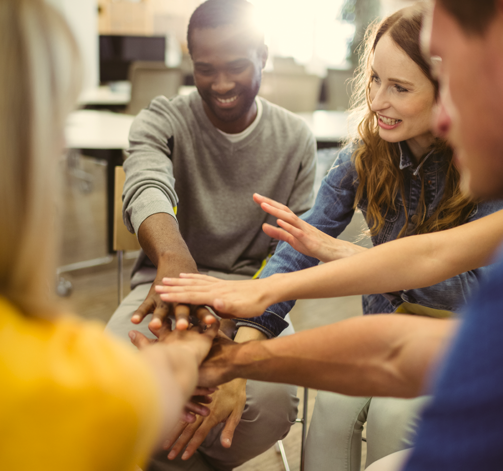 business-executives-with-hand-stacked 1 People gathered in a circle, stacking their hands in the middle to celebrate a shared achievement.