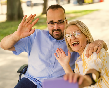 closeup-support-hands 1 (2) A man in a wheelchair waves at the camera while an older woman beside him poses playfully, both sharing a joyful moment.