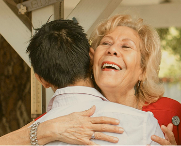 closeup-support-hands 1 A young man hugs an older woman in a warm front embrace, his back to the camera as she smiles with joy.