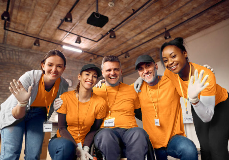 A cheerful team dressed in orange, with two members waving at the camera in an office setting. All are smiling and wearing work badges.