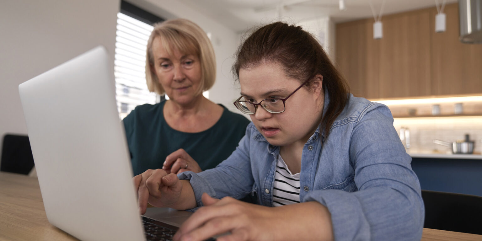 A girl with Down syndrome intensely staring at a laptop while an older woman supervises behind her.