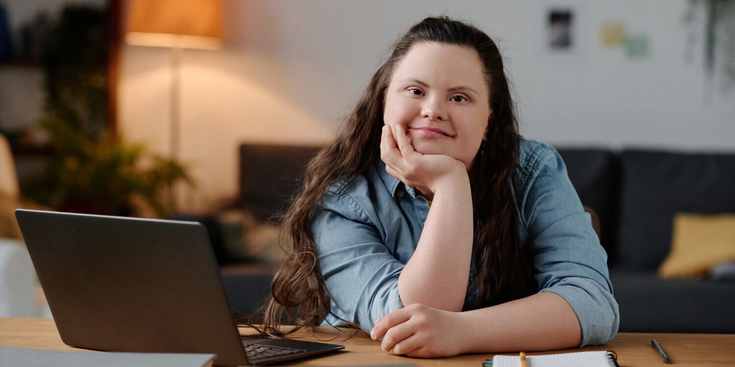 Girl with down syndrome studying at home A girl with Down syndrome smiling at the camera with a laptop in front of her.