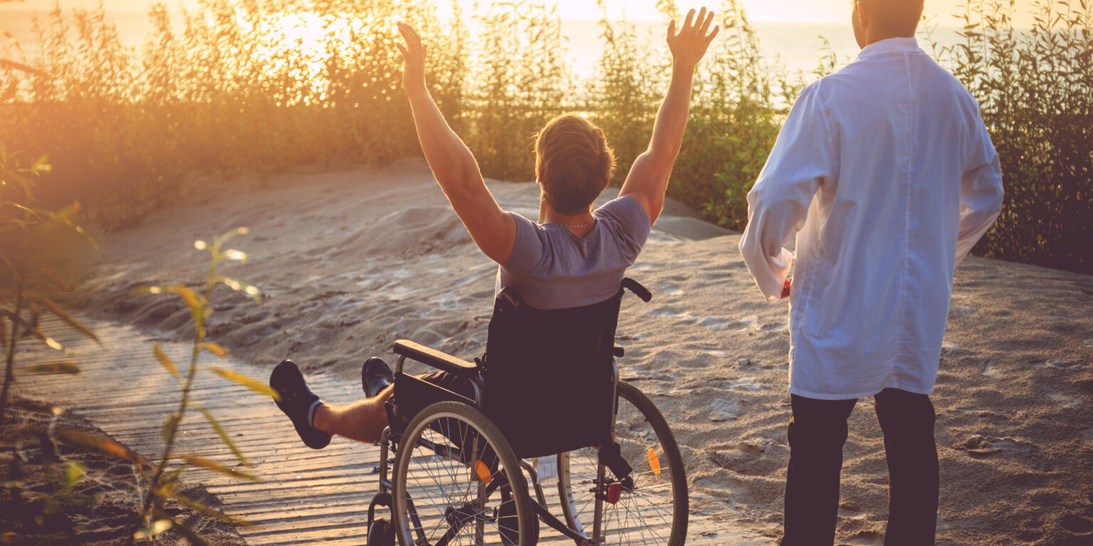 A man on wheelchair and his nurse enjoying sunrise on a beach.