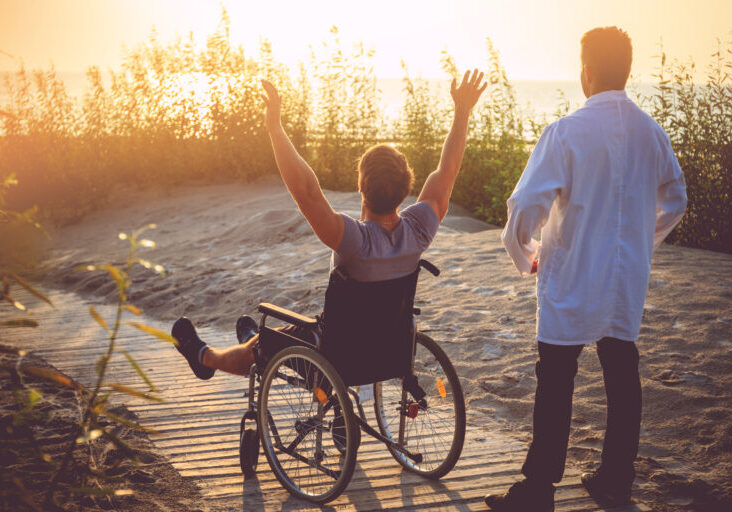 A man on wheelchair and his nurse enjoying sunrise on a beach.