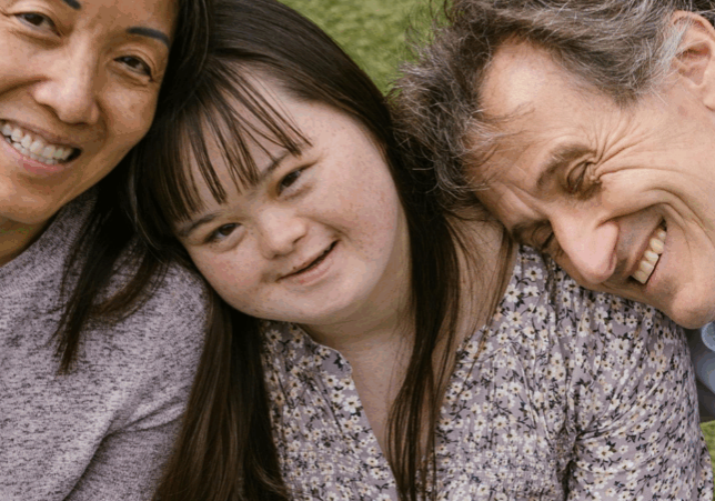 A young girl with Down syndrome smiling as a man rests his head on her shoulder and a woman rests her head on the opposite side of her head.