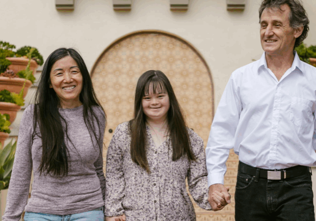 Two people holding the hands of a smiling girl with Down syndrome, one on each side.