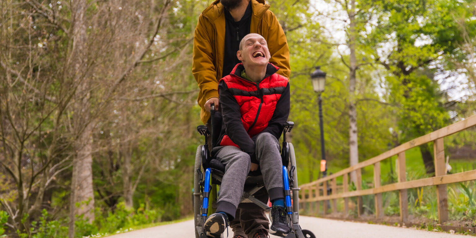 Paralyzed young man in the wheelchair being pushed by a friend in a public city park, strolling along a path having fun and smiling
