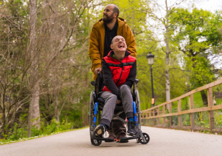 Paralyzed young man in the wheelchair being pushed by a friend in a public city park, strolling along a path having fun and smiling