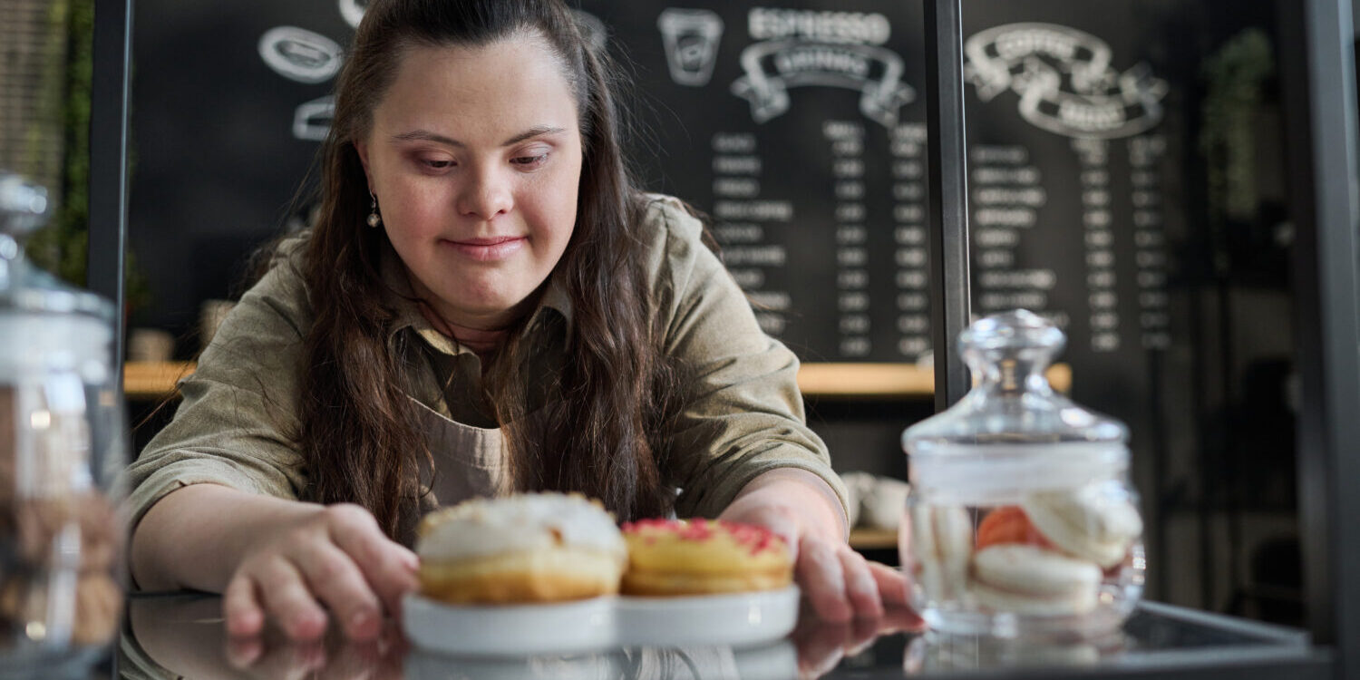 Waitress putting tasty donuts on tray Young smiling waitress with mental disability putting fresh tasty donuts on tray while preparing order of client by counter in front of camera